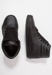 Para czarnych, wysokich sneakersów Vans z czarnymi sznurowadłami, widocznymi stawami i gumowymi podeszwami na białym tle.