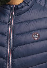 Marinefarbener Pufferjacke mit hohem Kragen, vertikal gestepptem Design und einem runden Logo-Patch. Verfügt über einen Reißverschluss mit roten und weißen Akzenten.