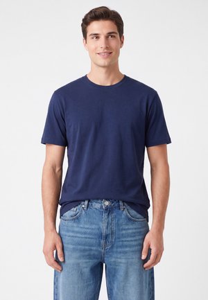 Jonge man draagt een effen marineblauw T-shirt met korte mouwen en lichtblauwe spijkerbroek, staand tegen een witte achtergrond.
