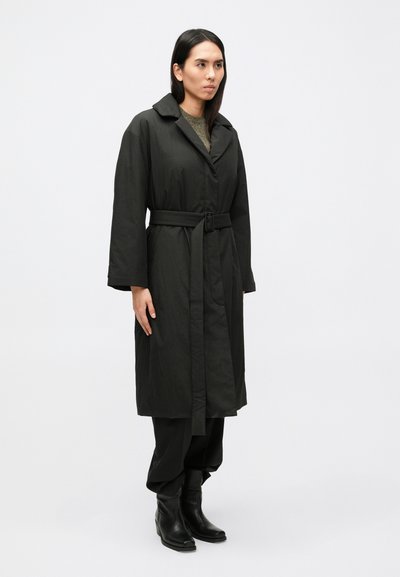 Čierny dlhý kabát s opaskom, so zárezovým golierom a širokými rukávmi. Nosený cez tmavé nohavice a členkové topánky. Hladká, matná textúra.