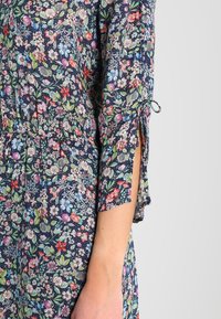 Kukkakuvioinen mekko, jossa tummansininen tausta, lyhyet hihat, joissa on nauhat, ja herkkiä kukkakuvioita eri väreissä, valmistettu kevyestä kankaasta.