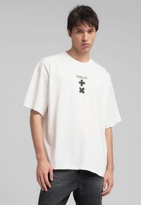 T-shirt bianco oversize in cotone con maniche corte. Presenta una grafica centrata con la scritta "REPLAY" e due simboli in nero. Design semplice e privo di decorazioni.