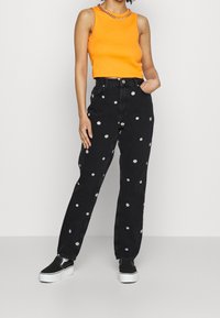 Oranžová bezrukávová blůza a černé džíny s vysokým pasem s bílými květinovými aplikacemi, doplněné černými nazouvacími botami a stříbrným řetízkem.