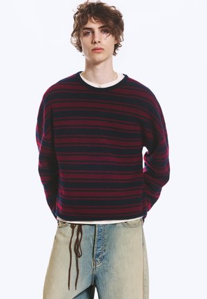 Junge Person mit lockigem Haar trägt einen dunkelblauen und rot gestreiften Pullover sowie hellblaue Jeans mit weitem Bein und sichtbaren Kordeln.