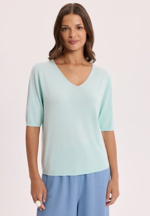 Kobieta o brązowych włosach do ramion, ubrana w jasnoniebieską bluzkę z dekoltem w serek i krótkim rękawem oraz luźne niebieskie spodnie na jednolitym tle.