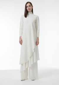 Witte lange mouwen jurk met hoge hals en asymmetrische zoom, gelaagd over wijduitlopende broek, gemaakt van een gladde stof, minimalistisch ontwerp.