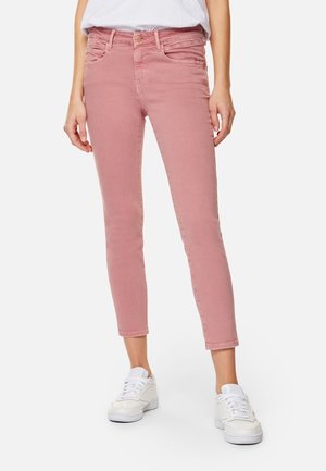 Žena v růžových těsně přiléhajících džínách s vysokým pasem a bílých teniskách stojí s rukama volně podél těla před jednobarevným pozadím.