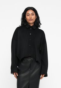 Čierna skrátená košeľa na gombíky s golierom, dlhými rukávmi a voľným strihom, skombinovaná s elegantnou čiernou koženou sukňou.