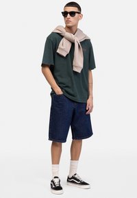 T-shirt en coton vert avec du texte rouge, sweat-shirt beige drapé autour du cou, short en denim bleu, baskets noires et chaussettes blanches.