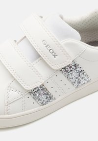 Baskets en cuir blanc avec deux bandes Velcro et des accents argentés scintillants. Comprend des perforations pour la respirabilité. Logo de la marque visible.