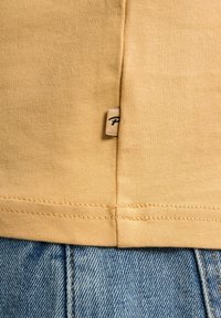 Un tricou din bumbac galben cu un eticheta cusută care prezintă un logo, așezat pe blugi de denim albastru. Materialul este neted, cu detalii de cusătură vizibile.