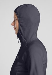 Giacca con cappuccio di colore grigio scuro, realizzata in materiale leggero e testurizzato. Presenta un design aderente e un cappuccio regolabile con dettagli arricciati.