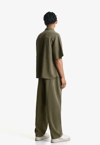 Cămașă verde măsliniu cu mânecă scurtă și pantaloni largi dintr-un material moale. Designul prezintă o croială lejeră și cusături vizibile.