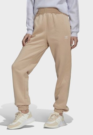 Onderlichaam van een persoon die beige Adidas trainingsbroek en witte sneakers draagt, staand met één been gekruist voor het andere.