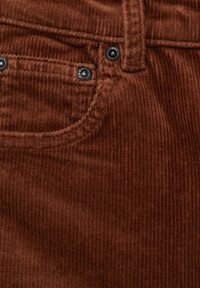 Gros plan sur un tissu en velours côtelé marron montrant une poche avant, une passants de ceinture et des boutons rivets en métal noir sur un pantalon texturé.