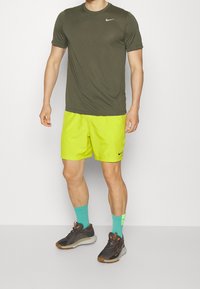 T-shirt verde oliva Nike, pantaloni corti giallo neon, calze turchesi e scarpe sportive nere e arancioni, che mostrano abbigliamento sportivo casual.