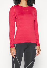 Punainen pitkähihaisella urheiluvaate, jossa on pyöreä kaula-aukko ja oranssit tikkausviivat, valmistettu kosteutta siirtävästä materiaalista. Yhdistetty mustiin leggingseihin.