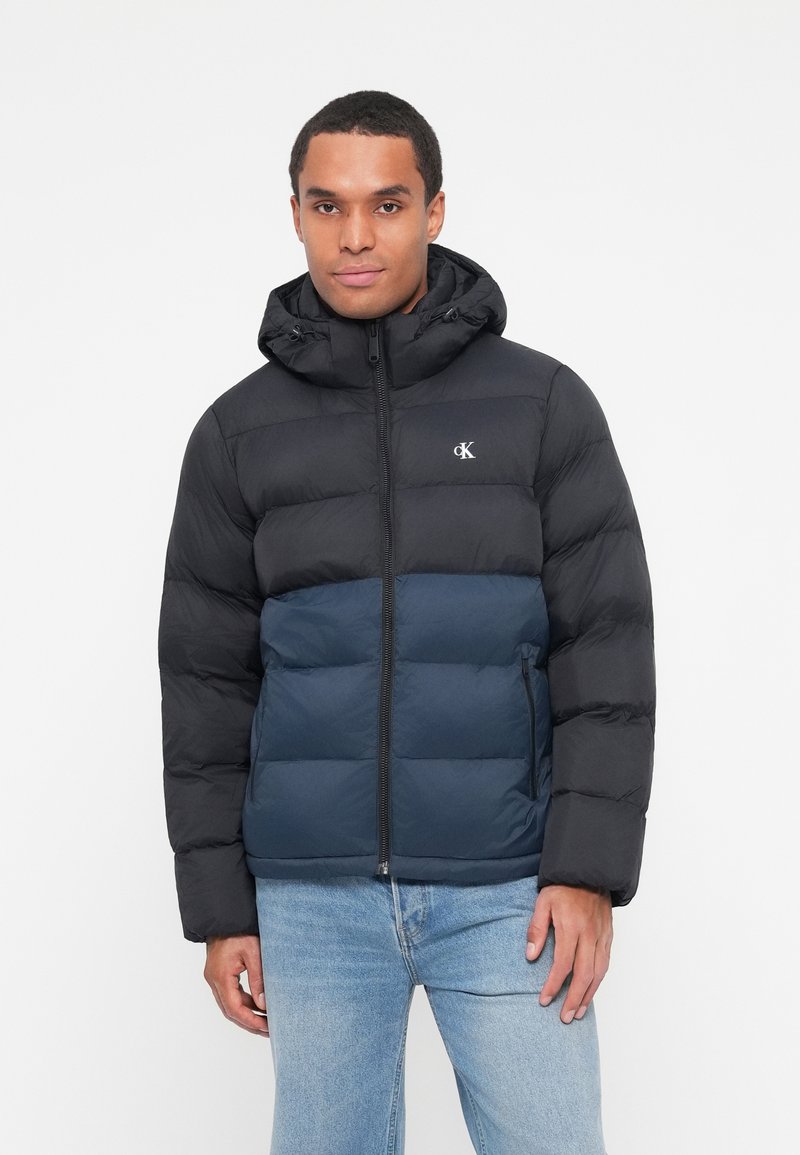 Padded bunda s dvoubarevným dizajnom v čiernej a tmavomodrej farbe. Obsahuje kapucňu, zips na zapínanie a logo na hrudi. Štruktúrovaná látka.
