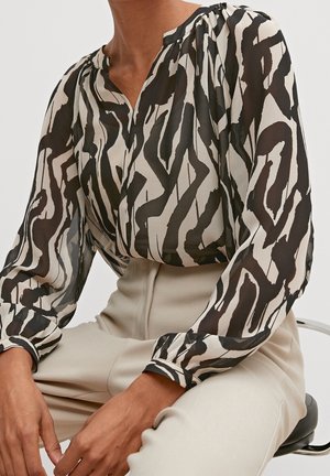 Kobieta ubrana w bluzkę z długim rękawem o czarno-białym abstrakcyjnym wzorze, włożoną do beżowych spodni z wysokim stanem, siedząca na stołku.