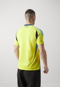 Żółta koszulka sportowa z niebieskimi akcentami, krótki rękaw i lekka, teksturowana tkanina. Napis "SVERIGE" na górze pleców.