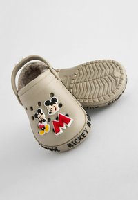 Beige-sinisiä klomppuja, jotka on valmistettu synteettisestä materiaalista, pörröisellä vuorilla, jossa on Mikki Hiiri -koristeita ja uritetut pohjat pidon parantamiseksi.