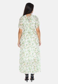 Robe maxi florale en vert clair avec une taille cintrée, des manches courtes bouffantes et un détail à ouverture en forme de clé dans le dos, associée à des chaussures à talons noires.