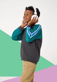 Junge trägt einen grauen Pullover mit türkisfarbenen Ärmeln und weißen Streifen, Khakihosen und weiße Kopfhörer, steht vor einem farbenfrohen Hintergrund.