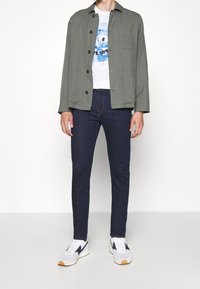 Olivově zelená bunda s černými knoflíky přes bílé tričko s potiskem. Tmavě modré přiléhavé džíny a bílé tenisky s námořnickými akcenty.