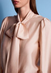 Zīda blūze maigi rozā tonī ar kakla mezglu, pūkainām piedurknēm un gludu tekstūru.