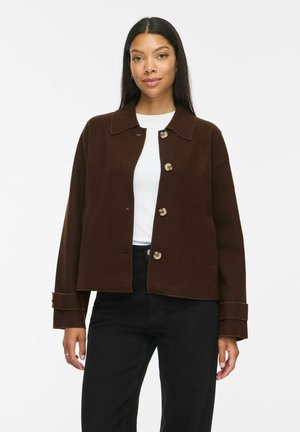 Femme portant une veste marron foncé boutonnée sur une chemise blanche et un pantalon noir, debout devant un fond blanc uni.