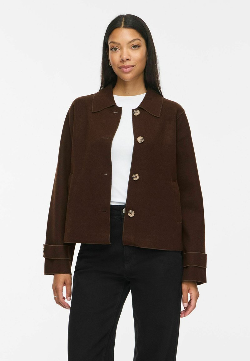 Femme portant une veste marron foncé boutonnée sur une chemise blanche et un pantalon noir, debout devant un fond blanc uni.