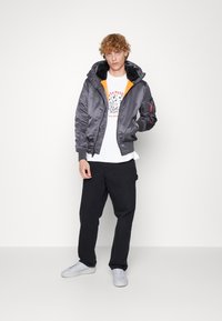 Gray bomber jacket with an orange lining, features a hood, ribbed cuffs, and a zip pocket on the sleeve, paired with a white graphic t-shirt and black pants.