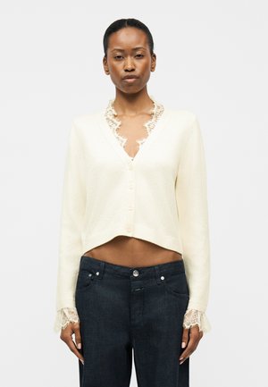 Kobieta ubrana w biały, krótki kardigan z koronką na kołnierzu i mankietach, w połączeniu z ciemnymi jeansami z wysokim stanem, stojąca na białym tle.