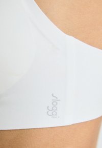 Hvit bh med sømløst design, myk tekstur og synlig merkevarelogo på bandet. Materialet ser ut til å være elastisk og figurnær.