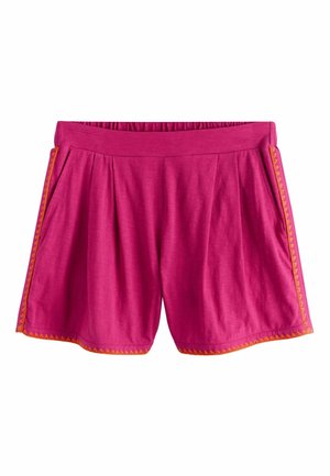 Frau mit schwarzen Haaren, die ein ärmelloses magentafarbenes Top und passende Shorts mit orangenem Rand trägt, steht an einem Sandstrand am Meer.