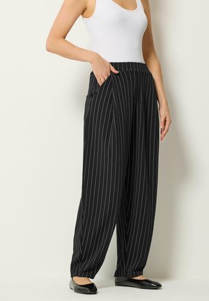 Frau trägt hoch taillierte, schwarze, breit geschnittene Anzughose mit Nadelstreifen, Hand in der Tasche, kombiniert mit einem weißen ärmellosen Oberteil und schwarzen Ballerinas.