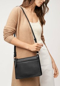 Sac à bandoulière en cuir noir avec une forme rectangulaire, une sangle ajustable et un design minimaliste ; il présente une texture lisse et des coutures discrètes.