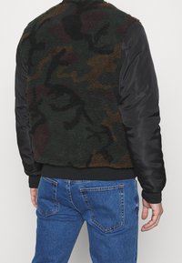 Man som bär en fleecejacka med kamouflage-mönster och svarta nylonärmar, tillsammans med blå denimbyxor, sedd bakifrån.