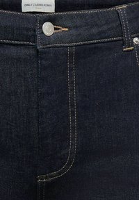 Tmavomodré džínsy z denimu v detailnom zábere ukazujúce predný gombík, slučky na opasok a jemný kontrastný šv s bielou nálepkou s nápisom "ONLY CARMAKOMA Denim."