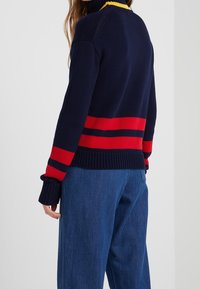 Personne portant un pull en maille bleu marine avec des rayures horizontales rouges et noires et des manches retroussées, associé à un jean denim bleu ample.