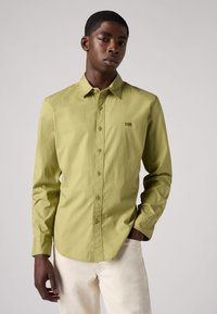 Camicia a maniche lunghe verde chiaro con colletto button-down, caratterizzata da una discreta toppa con logo e un tessuto liscio e testurizzato. Abbinata a jeans bianchi.