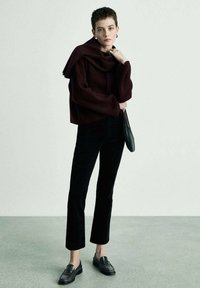 Burgundska sv sweater sa širokim izrezom, crne skraćene traperice i crne cipele bez pete. Manekenka drži malu crnu torbicu, stojeći na teksturiranom podu.