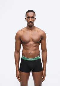 Μυώδης άνδρας που φοράει μαύρα εσώρουχα Lacoste boxer με πράσινη ζώνη, στέκεται μπροστά σε απλό λευκό φόντο, κοιτάζοντας προς τα εμπρός.