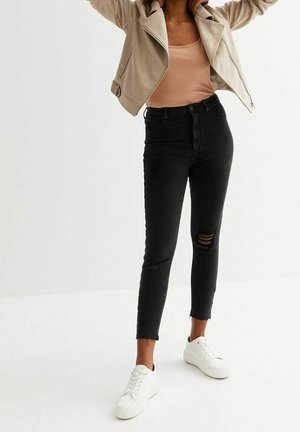 Person iført beige jakke, tan top, sorte revnede skinny jeans og hvide sneakers, der står mod hvid baggrund.