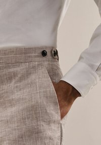 Ljusgrå rutiga byxor i en strukturerad textil med två svarta knappar i midjan och en sidoficka. En vit skjortärm syns.