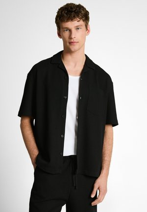 Młody mężczyzna o kręconych włosach, ubrany w czarną koszulę na guziki z krótkim rękawem na białym podkoszulku, stoi z rękami swobodnie opuszczonymi.