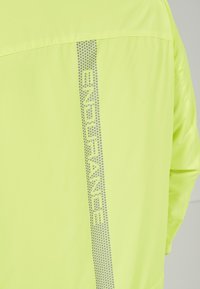 Veste fluorescente jaune avec une texture lisse. Présente un logo argenté réfléchissant "ENDURANCE" et un motif de rayures à pois dans le dos.