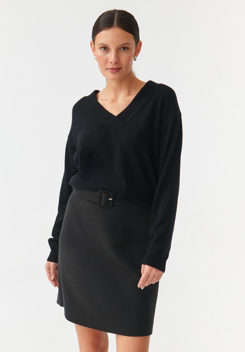Schwarzer V-Ausschnitt-Pullover mit langen Ärmeln, kombiniert mit einem taillierten schwarzen Rock, der einen Gürtel mit Schnalle hat. Beide Teile haben eine glatte Textur.