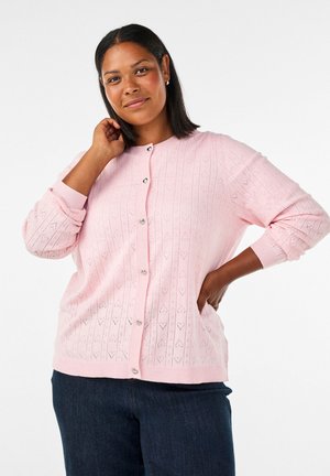 Vrouw met een gemiddelde huidtint en recht zwart haar, die een roze gebreide trui met knopen en hartpatronen draagt en een donkerblauwe jeans, staand met de hand op de heup.