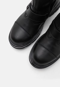 Bottes cheville en cuir noir avec embout renforcé, bride latérale, texture lisse et semelle en caoutchouc. Comprend des détails de couture et un accent en métal circulaire.